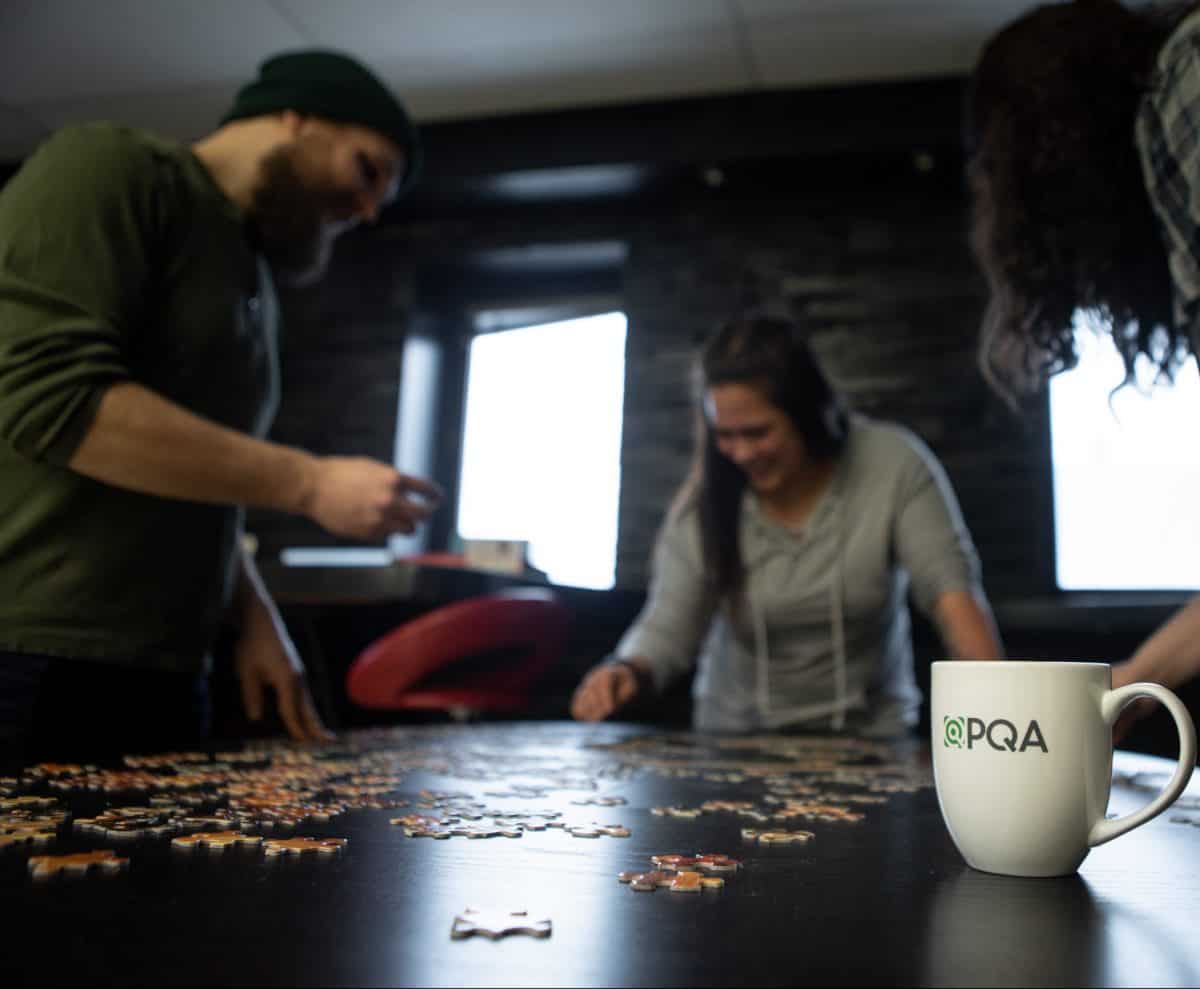 PQA coffee mug with employees in the background doing puzzle