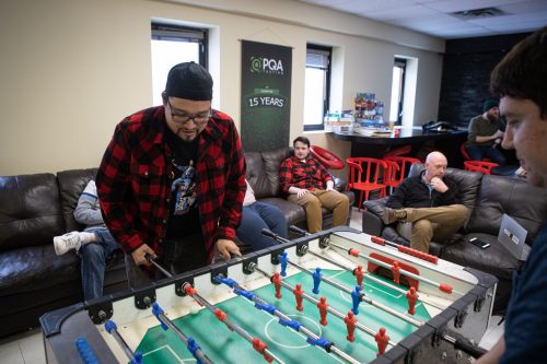 Two male PQA employees play fooseball while others talk behind them