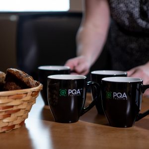 PQA coffee mugs being placed on a wooden table beside basket of muffins